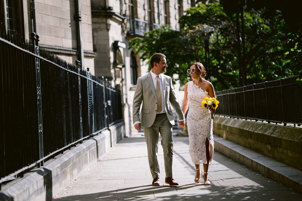 A bride in a white lace dress and a groom in a light suit walk hand-in-hand down a pavement beside a black iron fence during an intimate Sheffield wedding.