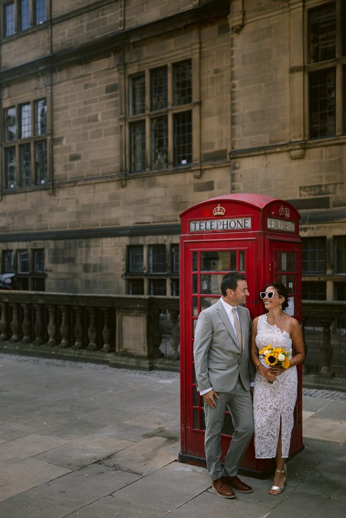 A bride and groom in wedding attire stand next to a red British telephone box during their Sheffield Town Hall wedding.