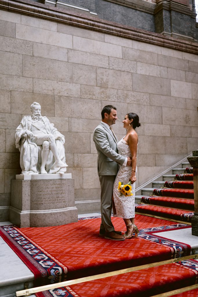A bride and groom stand face-to-face on the red carpeted staircase of Sheffield Town Hall, next to a large marble statue.
